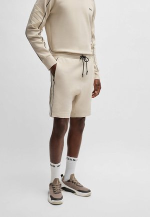Homme portant un sweat-shirt beige et un short avec un cordon noir, des chaussettes blanches et des baskets beiges, debout devant un fond uni.