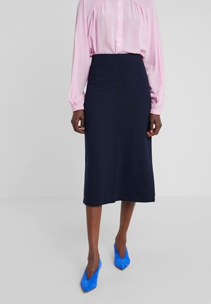 Femme portant un chemisier rose à manches longues, une jupe midi bleu marine, et des escarpins pointus d'un bleu vif, debout devant un fond uni.