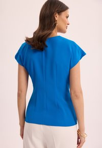 Dopasowana niebieska bluzka z krótkimi rękawami, gładka faktura i pionowe szwy podkreślające sylwetkę, w połączeniu z jasnymi spodniami.