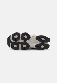 Talpa de cauciuc neagră cu un model texturat, având accente în formă de diamant. Mijloc de talpă de culoare gri deschis cu brandul New Balance.