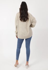 Suéter de punto beige de corte holgado con dobladillo acanalado, combinado con vaqueros ajustados azules y sandalias de tacón de bloque transparentes. Textura suave.