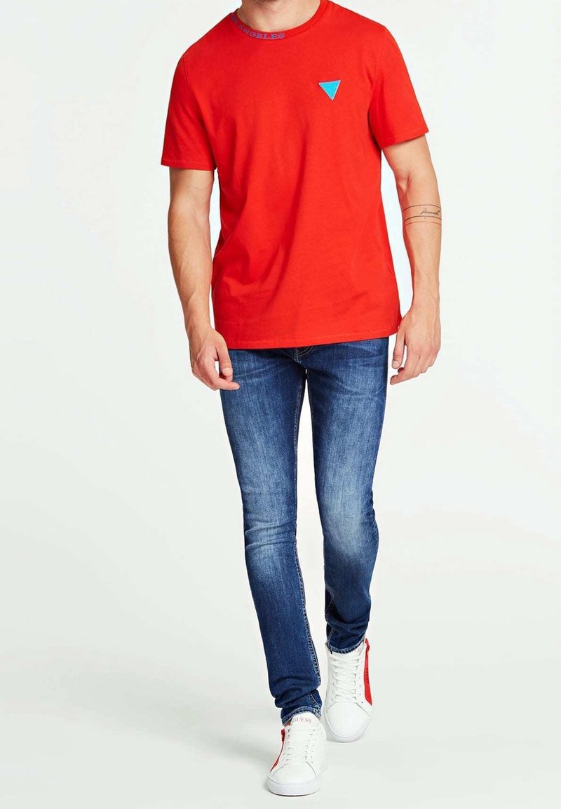T-shirt en coton rouge à col rond, avec un logo triangle bleu sur la poitrine ; associé à un jean skinny bleu et des baskets blanches.