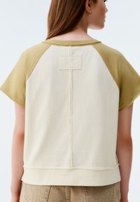 Short-sleeve top featuring a cream body with olive raglan sleeves, textured stitch detail along the seam, and a square patch on the back.