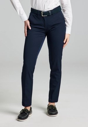 El modelo lleva pantalones azul marino con un cinturón negro, una camisa blanca y mocasines negros sobre un fondo gris liso.