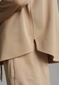 Haut beige à plusieurs couches avec une coupe décontractée, fabriqué en tissu doux. Détails notables : ourlet incurvé et accents de couture subtils.