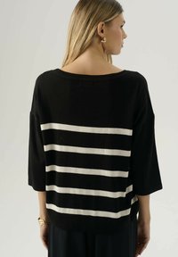 Donna di spalle con un maglione nero con quattro strisce bianche orizzontali sulla schiena e maniche a tre quarti.
