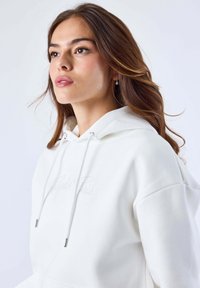 Felpa bianca con cappuccio regolabile, caratterizzata da un dettaglio in testo inciso. Tessuto morbido e vestibilità rilassata. Stoffa liscia con design minimalista.