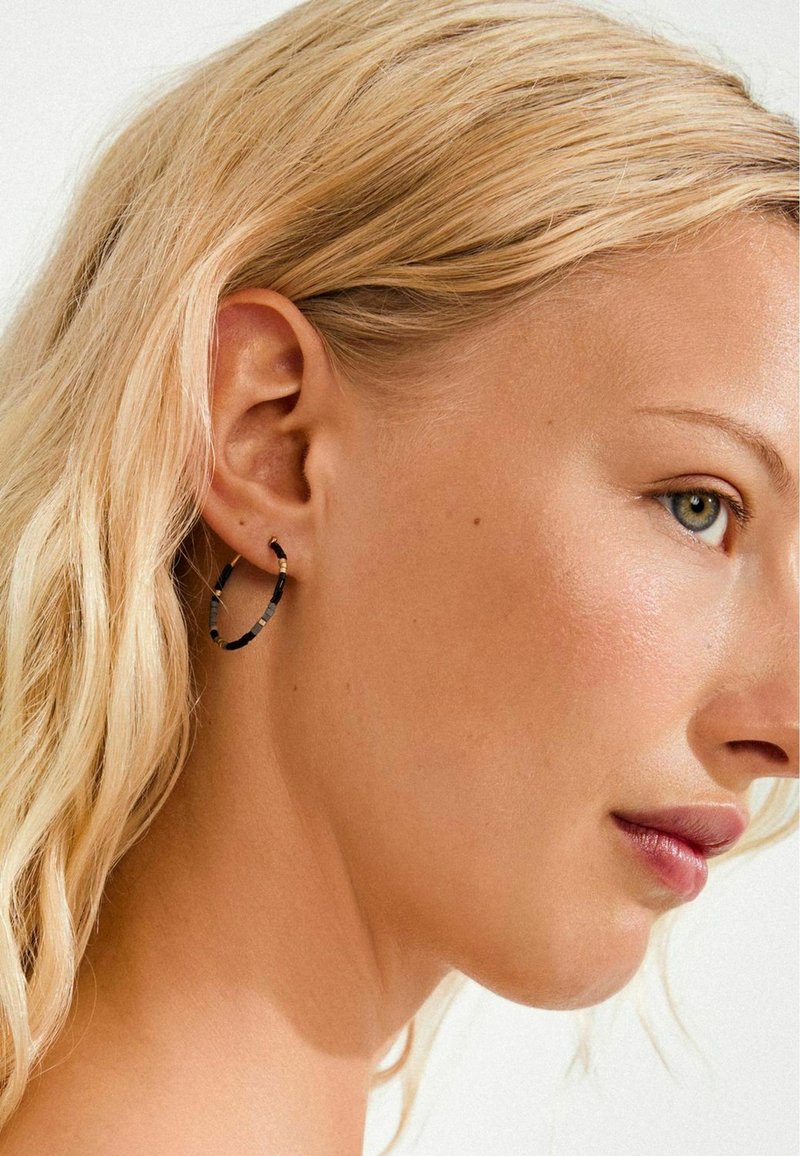 Boucles d'oreilles créoles avec un motif noir et or, finition métallique, forme circulaire, suspendues à l'oreille, présentant une texture lisse.
