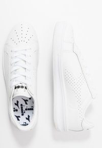 Sneakers bianchi con dettagli perforati, tomaia in pelle liscia, lacci piatti e suole in gomma. L'interno presenta un motivo a logo bianco e nero.