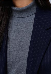 Harmaa korkeakauluksinen villapaita, jossa on sileä pinta, yhdistetty tummansiniharmaaseen raitapukuun. Villapaidassa on muotoiltu kaula-aukko ja hihat.