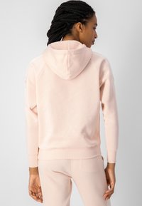 Sudadera con capucha de color rosa claro, con mangas largas, puños acanalados y un dobladillo elástico. Presenta una textura suave y un diseño minimalista.