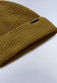 Gorro de punto color caqui con textura acanalada y puño doblado. Presenta una etiqueta de marca negra en el costado.