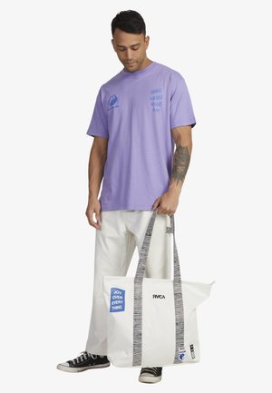 White tote bag with a blue rectangle reading "JOY OVER EVERYTHING," black striped handles, and a stylish black logo. Accompanied by a purple T-shirt.