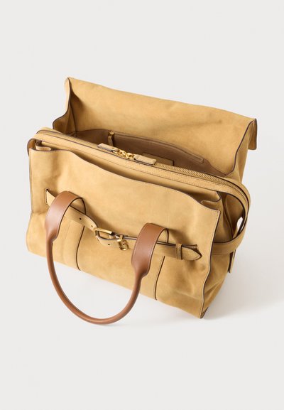Sac fourre-tout en daim couleur fauve avec anses en cuir marron, une sangle avant avec détail de boucle et un compartiment principal zippé partiellement ouvert.