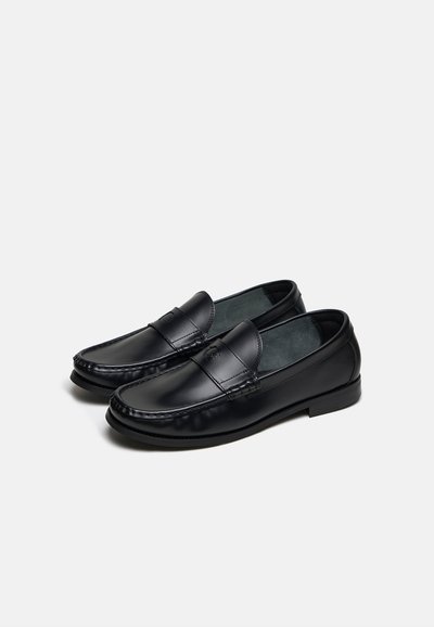 Ζευγάρι μαύρα δερμάτινα παπούτσια τύπου slip-on με ραμμένες λεπτομέρειες και ελαφρώς ανυψωμένη φτέρνα σε απλό ανοιχτό φόντο.