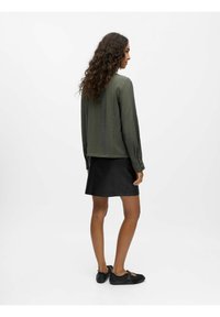 Olivenfarvet langærmet skjorte med tekstureret stof, parret med en sort nederdel. Outfittet er suppleret med sorte sko med stropper.