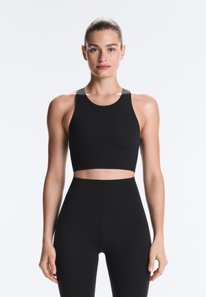 Jeune femme aux cheveux plaqués en arrière portant un crop top noir à col montant et des leggings taille haute assortis, debout devant un fond uni.