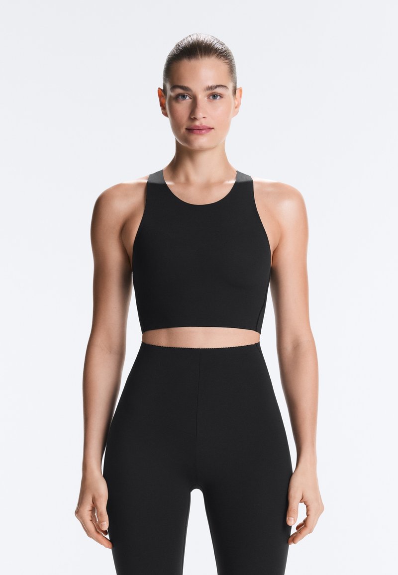 Jeune femme aux cheveux plaqués en arrière portant un crop top noir à col montant et des leggings taille haute assortis, debout devant un fond uni.