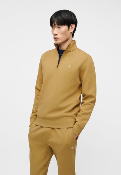 Νέος άνδρας που φοράει μπεζ φούτερ με φερμουάρ και ταιριαστό παντελόνι φόρμας, με τα χέρια στις τσέπες, κοιτάζοντας πλάγια μπροστά σε απλό φόντο.