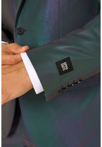 Donkergroene, iriserende manchetknoop van een colbert met een wit overhemd eronder. Voorzien van een zwart label met de tekst "OPPO SUITS" en een knoopsluiting.