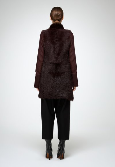 Donna con capelli castani raccolti in un ponytail, che indossa un gilet in pelliccia scura senza maniche, una camicia traslucida a maniche lunghe, pantaloni larghi neri e stivali neri con tacco alto.