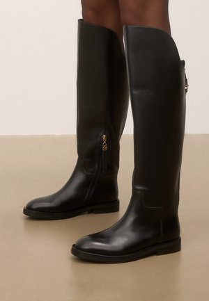 Bottes en cuir noires montantes jusqu'aux genoux avec une semelle plate et une fermeture éclair latérale portées sur des jambes brunes sur fond beige.