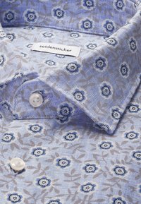 Chemise boutonnée bleu clair avec un petit motif floral bleu et gris, étiquette visible "seidensticker" à l'intérieur du col.