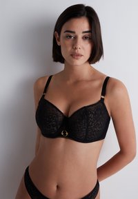 Soutien-gorge noir en dentelle avec armatures, présentant un motif floral, des bretelles fines et ajustables, ainsi qu'un accent en or au centre.