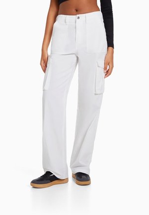Kvinde iført hvide cargobukser med høj talje, for- og sidelommer, kombineret med sorte sneakers med gummisåler.