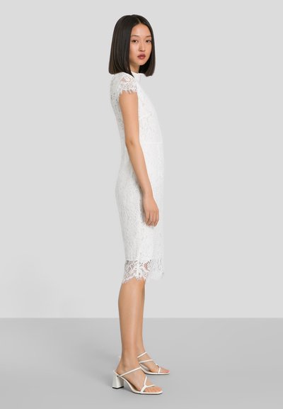 Λευκό φορεματάκι δαντέλας με κοντά μανίκια, εφαρμοστή σιλουέτα και λεπτομέρειες δαντέλας στη βάση. Φορεμένο με λευκές σανδάλες με μπλοκ τακούνια.