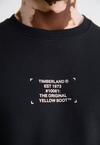 Černá mikina s kulatým výstřihem a bílým textem: "TIMBERLAND EST 1973 #10061: PŮVODNÍ ŽLUTÁ BOOT™" v rámovaném designu.