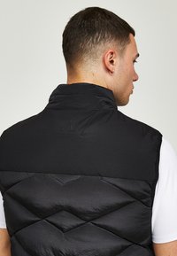 Črn napihnjen brezrokavnik z visokim ovratnikom, quilted oblikovanjem in teksturirano tkanino. Diskretno blagovno znamko na hrbtni strani, prešit čez belo majico.