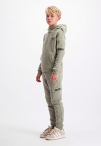 Blond kind staat in een olijfgroene hoodie en bijpassende cargobroek met beige sneakers tegen een witte achtergrond.
