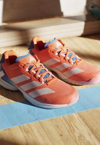 Coppia di scarpe da corsa atletiche arancioni e bianche con foderina interna blu, posizionate su un pavimento in legno con una linea blu dipinta.