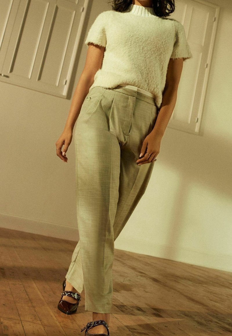 Personne portant un haut blanc duveteux à manches courtes, un pantalon beige taille haute à jambes larges et des ballerines foncées avec brides à la cheville, debout sur un sol en bois.