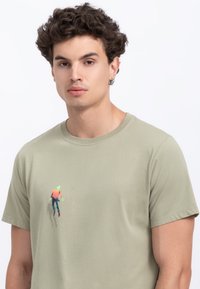 Olijfgroene katoenen t-shirt met een ronde hals en korte mouwen, met een kleine levendige print van een figuur in kleurrijke kleding op de borst.