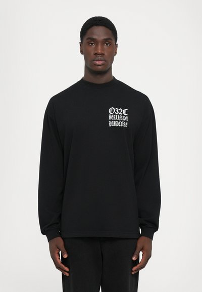 Mann trägt ein schwarzes Langarmshirt mit weißem gotischen Text "032C BERLIN 2001 HARDCORE" auf der linken Brust und steht vor einem schlichten weißen Hintergrund.
