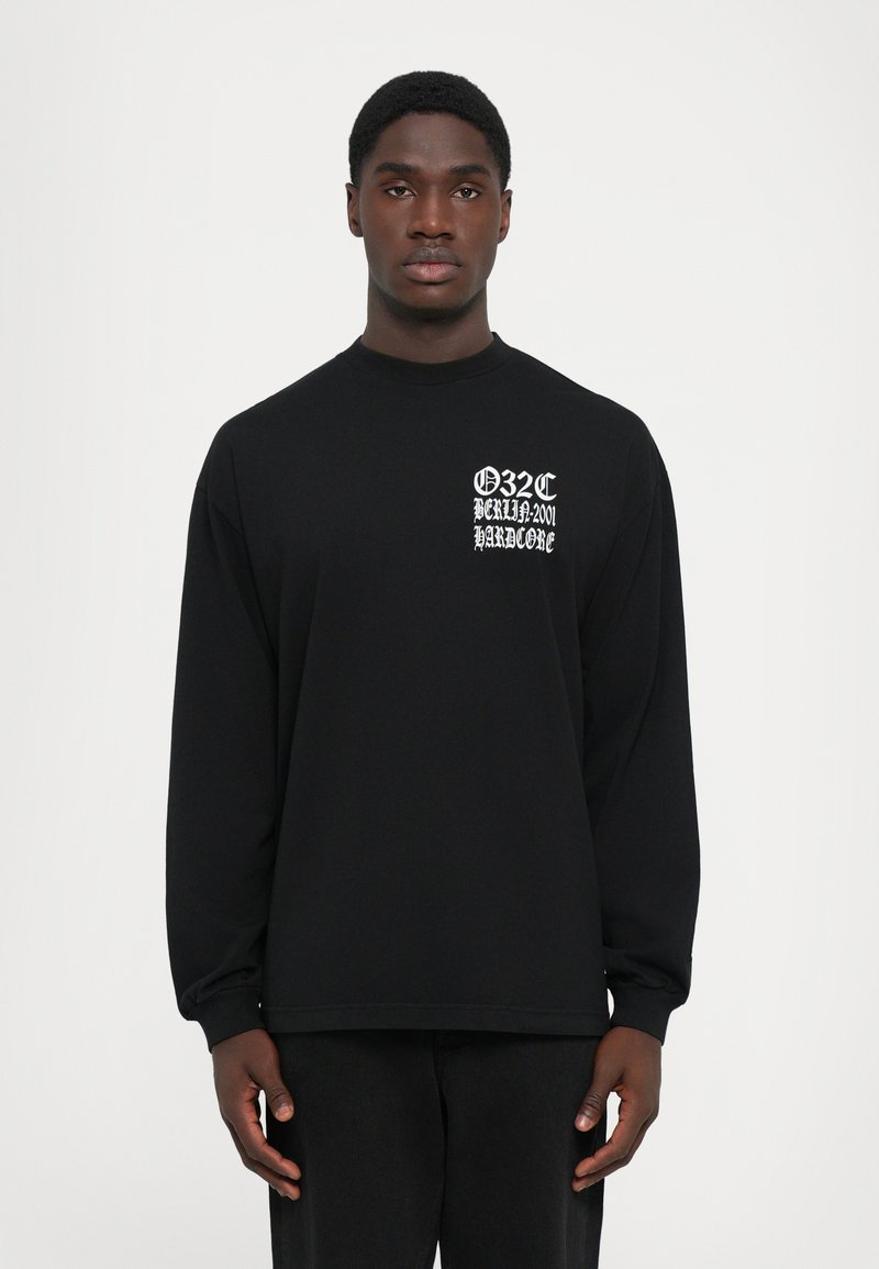 Homme portant une chemise à manches longues noire avec un texte gothique blanc "032C BERLIN 2001 HARDCORE" sur la poitrine gauche, se tenant devant un fond blanc uni.