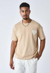 Camiseta de manga corta beige con un diseño de corazón texturizado en el lado izquierdo del pecho, combinada con pantalones blancos. Tejido suave, corte relajado.