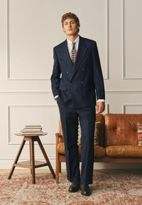 Jeune homme en costume croisé bleu marine, cravate à motifs, chemise blanche, chaussures noires, debout à côté d'une table d'appoint en bois avec des livres et un canapé en cuir fauve.