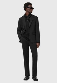 Sort skræddersyet jakkesæt med knaplukket blazer, lynlås krave og tætsiddende bukser. Struktureret stof med glat finish, båret med sorte solbriller.