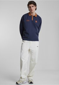 Navy langermet polo skjorte med brun krage, med broderte tekst. Kombinert med hvite løstsittende bukser og lette sneakers.