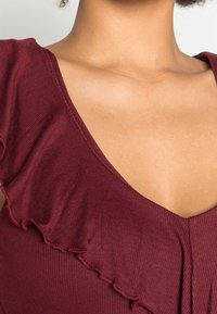 Top bordeaux con una texture a coste, caratterizzato da un profondo scollo a V e dettagli arricciati lungo il colletto e le spalle.