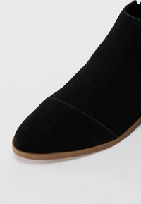 Prim-plan al unei cizme negre din suede cu vârful ascuțit și talpă de un maro deschis, pe un fundal alb.