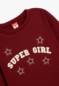 Vihreä puuvillapaita, jossa on valkoiset "SUPER GIRL" -tekstit ja tähtigrafiikat, joissa on ympyröityjä tähtiä ja sileä pinta.