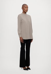 Lys beige langermet bluse med høy krage og elastiske mansjetter, kombinert med høyt liv svarte bukser og spisse svarte hæler.