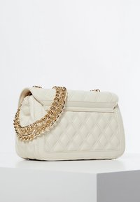 Nahkanahkainen, tikattu kermah-colored käsilaukku, jossa on kullanvärinen ketjuhihna ja suorakulmainen muoto. Huomionarvoinen geometrisen kuvion ja jäsennellyn rakenteen ansiosta.