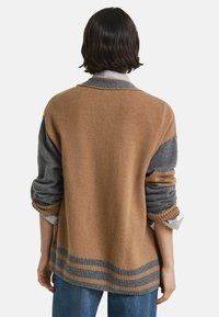 Personne aux cheveux foncés portant un pull bleu et marron à blocs de couleur, retroussé sur une chemise rayée, de dos contre un fond blanc.
