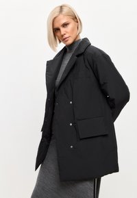 Chaqueta negra impermeable de corte relajado, con un gran bolsillo frontal, botones a presión y cuello de chal. Textura suave y lisa.