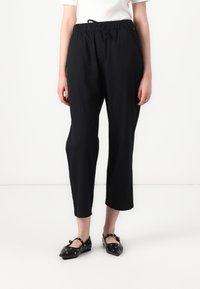 Pantalons noirs légers avec une taille élastique et un cordon de serrage. Longueur crop, texture lisse, sans motifs visibles, associés à des chaussures noires plates.